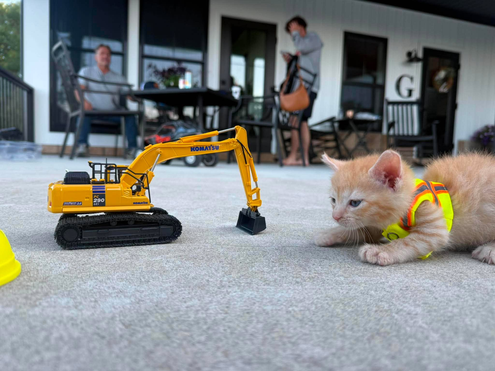 New Acts ($SUE) on Onchain Circus Sue in her hi-vis vest crouched nose to nose with a model Komatsu PC290 excavator, two people out of focus on the porch behind her.